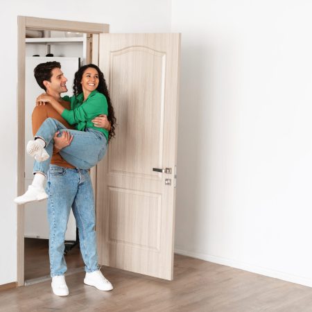 Loving millennial husband carrying wife in his arms standing in doorway, newlyweds celebrating moving day, happy family having fun in empty room, couple excited by relocation into first new house